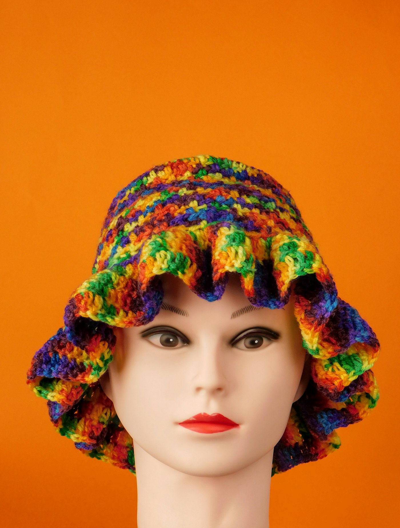 A rainbow crocheted bucket hat with a ruffled brim on an orange background